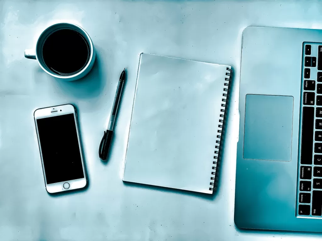Mesa com alguns objetos( caneta, celular, notebook, caneca de café e um caderno)